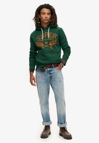 Sudadera verde con logotipo naranja, jeans azules desgastados y botas marrones. Diseño con capucha, estampado gráfico, puños enrollados en los jeans.