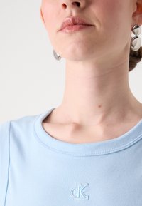 Světle modrý bavlněný top s kulatým výstřihem, zdobený vyšívaným logem "CK" na hrudi, doplněný stříbrnými kruhovými náušnicemi.