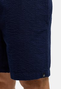 Pantalones cortos texturizados de color azul marino, confeccionados con tela tejida. Presentan un patrón sutil, bolsillos laterales y una pequeña etiqueta con el logo en el dobladillo.