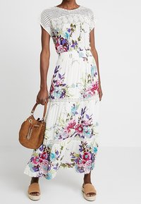 Maxi kjole i hvidt stof med blomsterdesign i pink og blå, blondekant ved udskæringen og taljen, parret med en brun taske og sandaler.