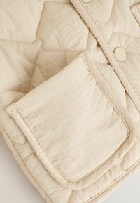 Beige gesteppte Weste mit Polsterung, ausgestattet mit einer Fronttasche und einer Knopfleiste. Weicher, strukturierter Stoff mit kontrastierenden Nähtelementen.