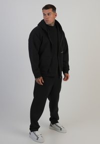 Melns kapuce ar rāvējslēdzēju, kas piedāvā lielas kabatas un brīvu piegriezumu, kombinēts ar atbilstošām melnām sporta biksēm. Baltas kedas piešķir kontrastu.