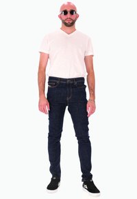 Jean en denim bleu foncé, coupe ajustée, associé à un t-shirt blanc à col en V. Baskets noires avec des accents blancs. Le porteur a la tête rasée et porte des lunettes de soleil.