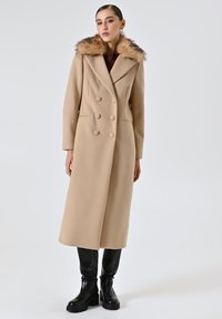 Cappotto doppiopetto in misto lana beige con colletto in pelliccia, dotato di due tasche laterali e dettagli in bottone. Abbinato a pantaloni neri aderenti e stivali.