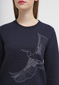 Navy quilted sweatshirt featuring a detailed white embroidered bird design on the front, round neckline, and textured fabric.