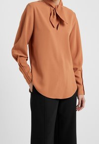 Blouse à manches longues couleur rouille avec un col à nouer, une patte de boutonnage à l'avant et un ourlet arrondi. Le tissu semble lisse et léger.