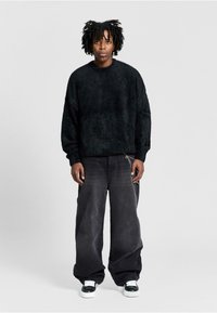 Schwarzer, flauschiger Pullover mit Rundhalsausschnitt, kombiniert mit lockeren, dunkelgrauen Jeans und schwarzen Sneakers mit weißen Schnürsenkeln und Akzenten.