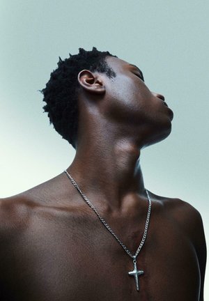 Rövid göndör hajú fiatal férfi, mezítelen mellkassal, ezüst kereszt nyakláncot visel, felfelé néz egy egyszerű világos háttér előtt.