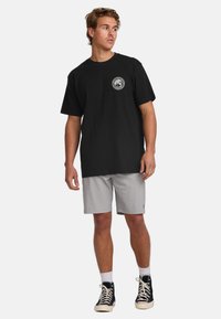 Black cotton t-shirt with a round graphic logo on the chest, paired with light gray shorts and black high-top sneakers.