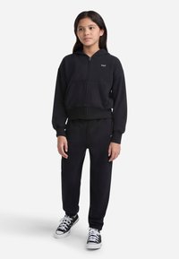 Sudadera corta con cremallera negra y bolsillos frontales, combinada con pantalones jogger a juego. Completada con unas zapatillas negras y blancas.