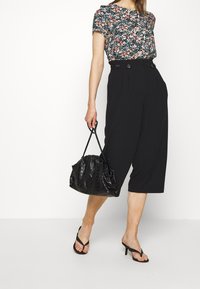 Bloemige top met korte mouwen op een donkere achtergrond, gecombineerd met hoge zwarte culottes. Het model draagt een gestructureerde zwarte handtas en zwarte sleehakken.