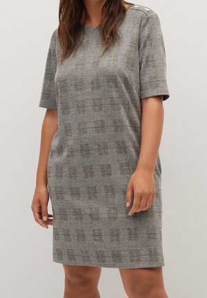 Kvinde iført en grå ternet kortærmet kjole med knapdetaljer på skulderen, stående mod en ensartet lys baggrund.