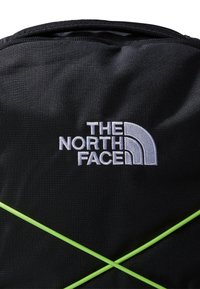 Mustat kangaskassit, joissa on selvästi näkyvä valkoinen The North Face -logo ja ristiin menevät vihreät hihnat etuosassa.