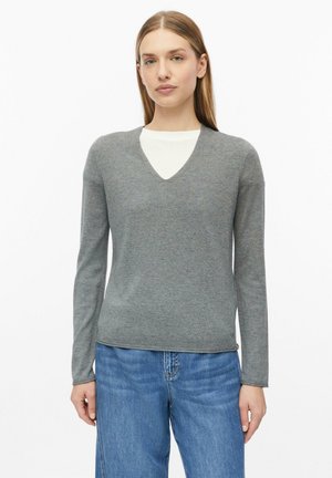 Femme aux cheveux lisses châtain clair porte un pull gris à col en V par-dessus un top blanc et un jean bleu, debout devant un fond blanc uni.