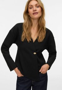 Juoda palaidinė su užrišamu stiliumi ir ilgomis rankovėmis, papuošta auksinio mygtuko detale. Pagaminta iš lygaus audinio, suteikia laisvą prigludimą ir modernų dizainą.