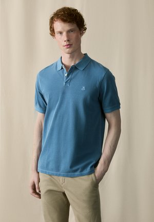 Hellblaues Polohemd mit einer Drei-Knopf-Leiste, kurzen Ärmeln und einem kleinen Logo auf der linken Brust. Getragen mit khakifarbenen Hosen.