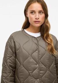 Jeune femme aux longs cheveux bruns portant une veste matelassée taupe sur une chemise blanche, regardant légèrement sur le côté sur un fond blanc.