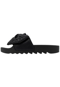 Black slide sandal with jagged sole and wide strap featuring a large decorative knot, isolated on white background.