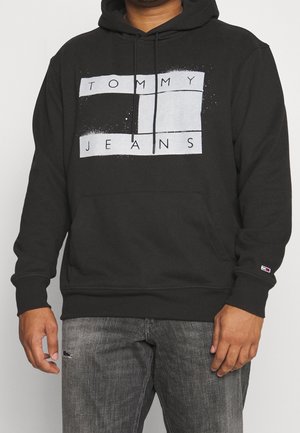Mann trägt schwarzen Hoodie mit weißem "TOMMY JEANS" Block-Logo und ausgewaschene graue Jeans mit kleinem Riss am linken Oberschenkel.