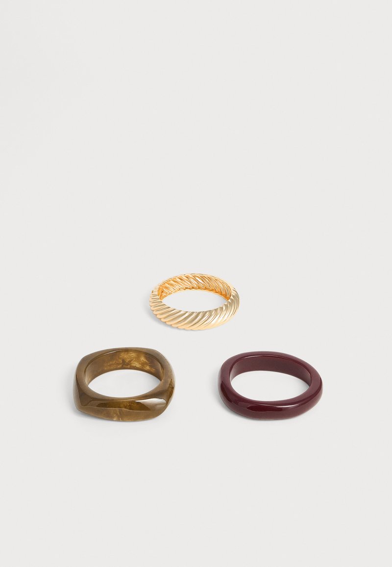 Trois bagues sur une surface blanche : une bande torsadée en or, une bague ronde rouge foncé brillante et une bague carrée marron brillante.