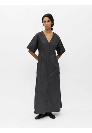 Femme debout devant un fond blanc, portant une longue robe noire à col en V avec des manches courtes et des chaussures plates noires, les mains derrière le dos.