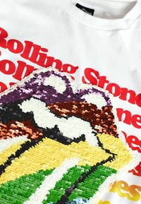 Valkoinen t-paita, jossa on monivärinen paljettivitsi kielen logolla, yläpuolella rohkeasti punaisella tekstillä lukee "Rolling Stones".
