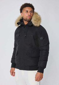 Doudoune noire avec capuche bordée de fourrure, poches zippées et poignets élastiques. Elle présente un extérieur chaud et lisse ainsi qu'une coupe décontractée.