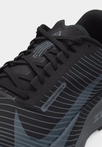 Black and gray athletic shoe with black laces, horizontal stripe pattern, and a label reading "All Conditions Gear" on the side.