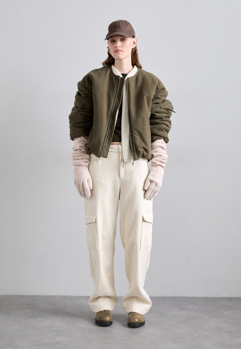 Olive green bomber jacket with a cream trim, beige textured pants with side pockets, light gloves, and a brown cap.