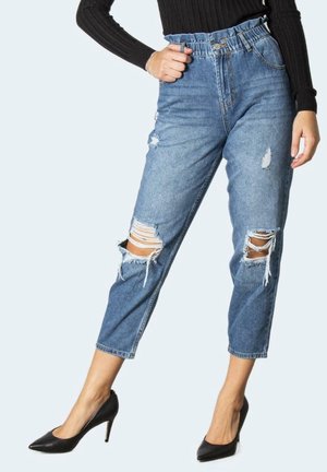 Mujer con jeans azules rasgados y tacones altos negros, con una mano apoyada en la cintura de los jeans.