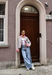 Šedá a růžová zkrácená bunda přes bílý top, spárovaná s volnými modrými džíny a bílými teniskami, na pozadí dřevěných dveří.