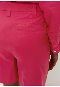 Pantalones cortos rosa brillante hechos de tela ligera. Presentan una textura suave, lazos para cinturón y un bolsillo trasero con costura horizontal.