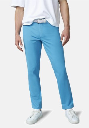 Mann trägt hellblaue, eng anliegende Hose, weißen Gürtel, weißes T-Shirt und weiße Turnschuhe, steht vor einem einfarbigen Hintergrund.