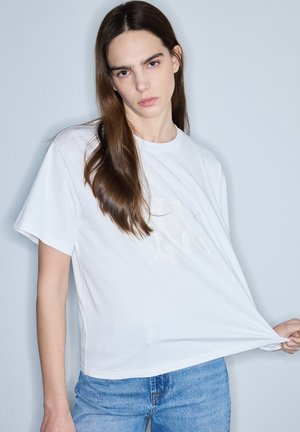 Frau mit langen braunen Haaren, die ein weißes T-Shirt und blaue Jeans trägt, zieht das Shirt mit einer Hand nach außen vor einem einfarbigen hellen Hintergrund.