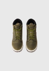 Une paire de bottes de randonnée en suède vert olive avec des lacets jaunes et noirs et des semelles blanches, vues de dessus sur un fond blanc.