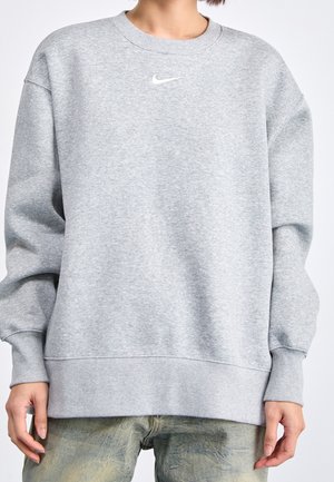 Osoba ubrana w jasnoszarą bluzę Nike z małym białym logo swoosh na środku klatki piersiowej, w połączeniu z wyblakłymi zielonymi spodniami.