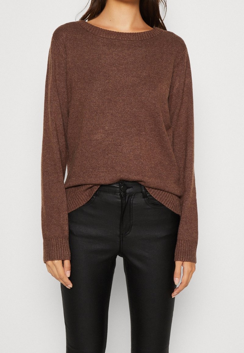 Femme portant un pull en maille brun à manches longues, légèrement rentré dans un pantalon noir ajusté taille haute, debout devant un fond uni.