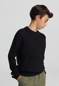 Czarny sweter dzianinowy o prążkowanej fakturze, okrągłym dekolcie i luźnym kroju, w połączeniu z oliwkowo-zielonymi spodniami.