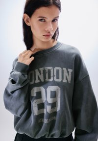 Harmaa ylipitkä collegepaita resoreilla hihoissa, jossa on etupuolella kuluneen valkoisella fontilla painettu teksti "LONDON 29 ATH.SEASON".
