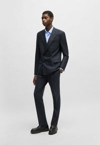 Dark navy double-breasted suit with structured shoulders, paired with a light blue checked shirt and black loafers. Smooth fabric texture.