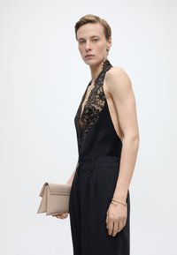 Γυναίκα με μαύρο τοπ από δαντέλα halter και ψηλόμεσο παντελόνι, κρατώντας μπεζ τσαντάκι, φορώντας χρυσά σκουλαρίκια και βραχιόλια, στέκεται μπροστά σε λευκό φόντο.