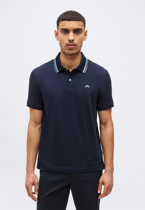 Ung mand med kort hår iført en marineblå polo med lyseblå stribet krave og et lille hvidt logo på brystet.