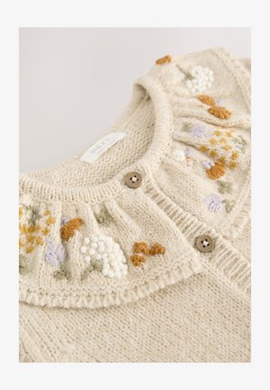 Kreminės spalvos megztinis su siuvinėtomis gėlių dekoracijomis gelsvos, violetinės ir žalios spalvų, su apvalia apykakle ir dviem rudais mygtukais.