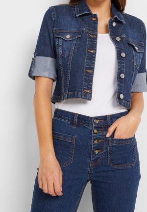 Korte spijkerjack met zilveren knopen, borstzakken en opgerolde mouwen. Gecombineerd met high-waisted jeans met knoopsluiting en zakken.
