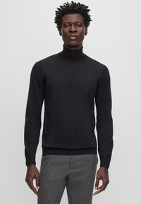 Pull à col roulé noir en tissu lisse, coupe ajustée, manches longues et poignets côtes, associé à un pantalon gris texturé.