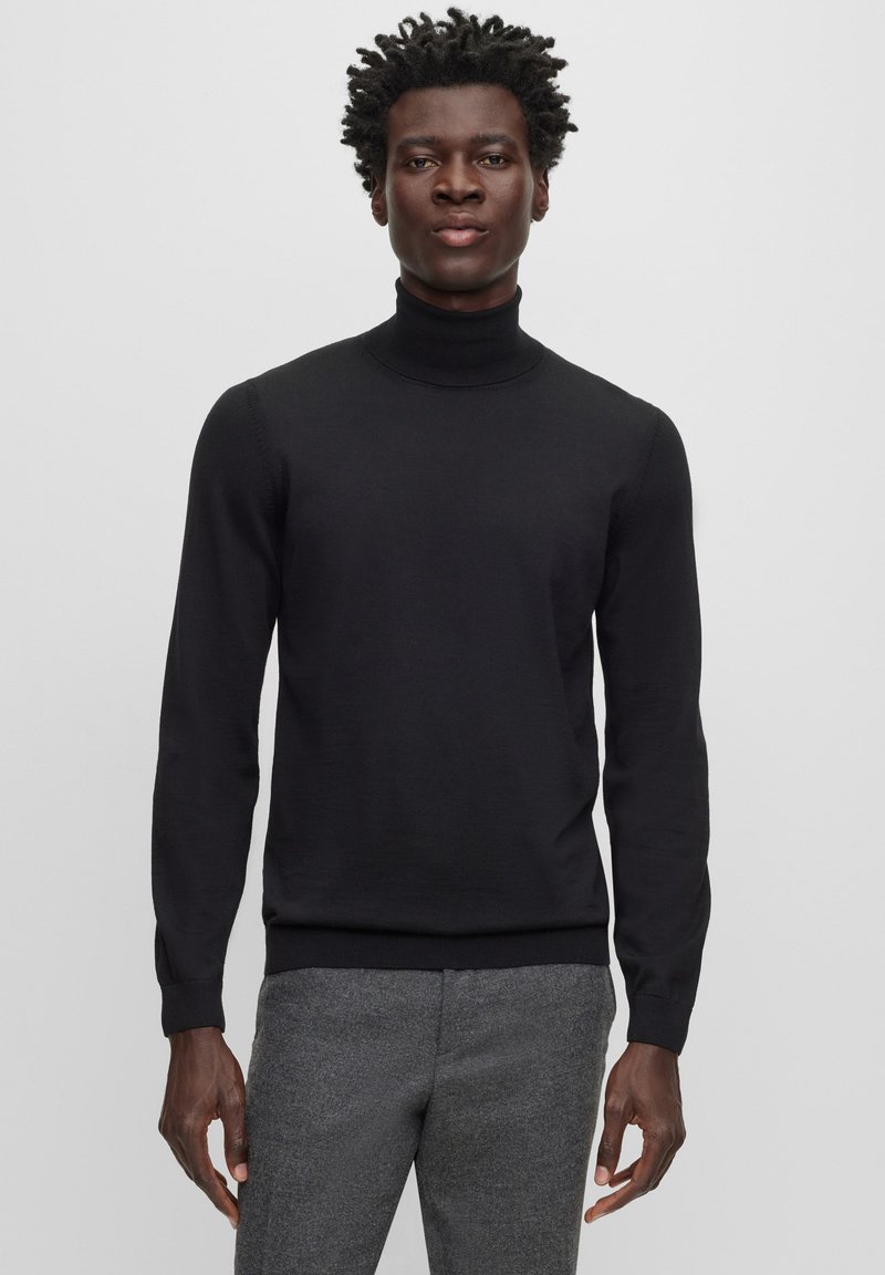 Pull à col roulé noir en tissu lisse, coupe ajustée, manches longues et poignets côtes, associé à un pantalon gris texturé.
