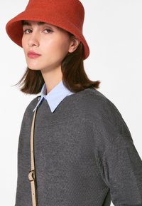 Grauer gestrickter Pullover mit strukturiertem Muster, darunter ein hellblaues Kragenshirt und ein orangefarbener Filzhut im Bucket-Stil. Langes braunes Haar ist teilweise sichtbar.