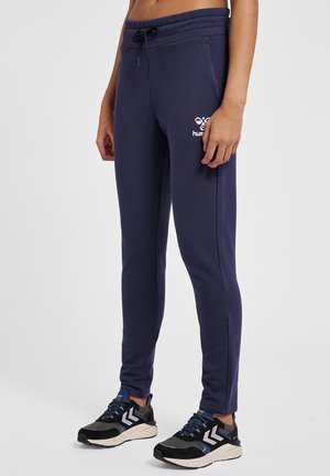 Kobieta ubrana w granatowe, dopasowane spodnie dresowe z logo na udzie, połączona z czarno-niebieskimi sportowymi butami na jednolitym tle.