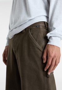 Olivgröna byxor med en texturerad tyg, sidofickor och en vävd etikett på midjan. Stylerade med en ljusgrå långärmad topp.