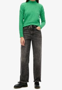 Pull en tricot vert avec un col montant, associé à un jean en denim noir à coupe droite. Des chaussures noires à lacets complètent la tenue.